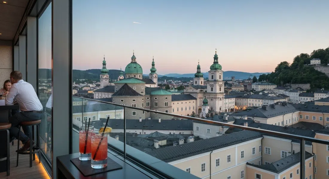 the glass-walled rooftop bar hu:goes14 on the 14th floor, cocktails in the foreground and Salzburg Old Town panorama at dusk