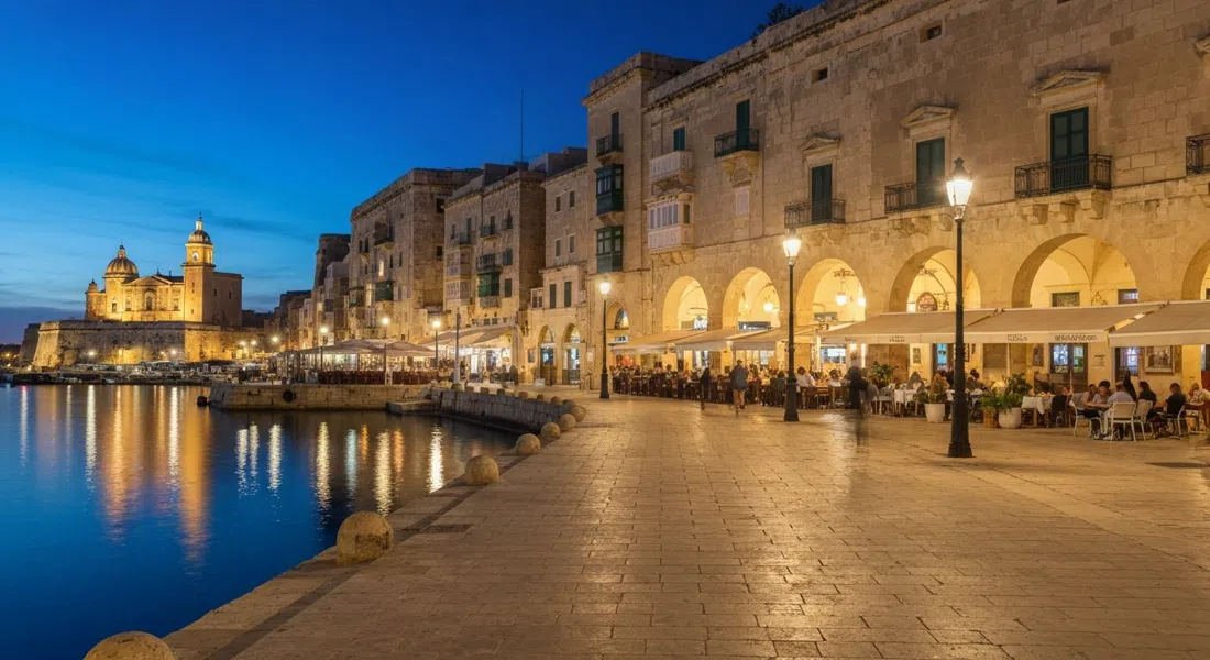 the Valletta Waterfront promenade at night with restaurant terraces lit under the arches and Grand Harbour reflections shimmering below the bastions