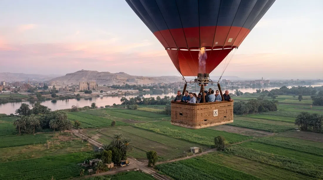 a sunrise hot-air balloon from Sindbad Balloons lifting above the West Bank fields, with the Nile and temple landscape beneath a pale pink sky