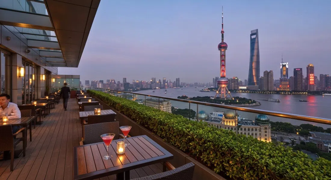 the open-air terrace at Flair on the 58th floor of the Ritz-Carlton Pudong at dusk, cocktails on tables and the Oriental Pearl Tower glowing across the river